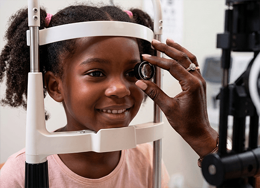 A young girl at the examination frame, smiling, while a lens is held up to her eye